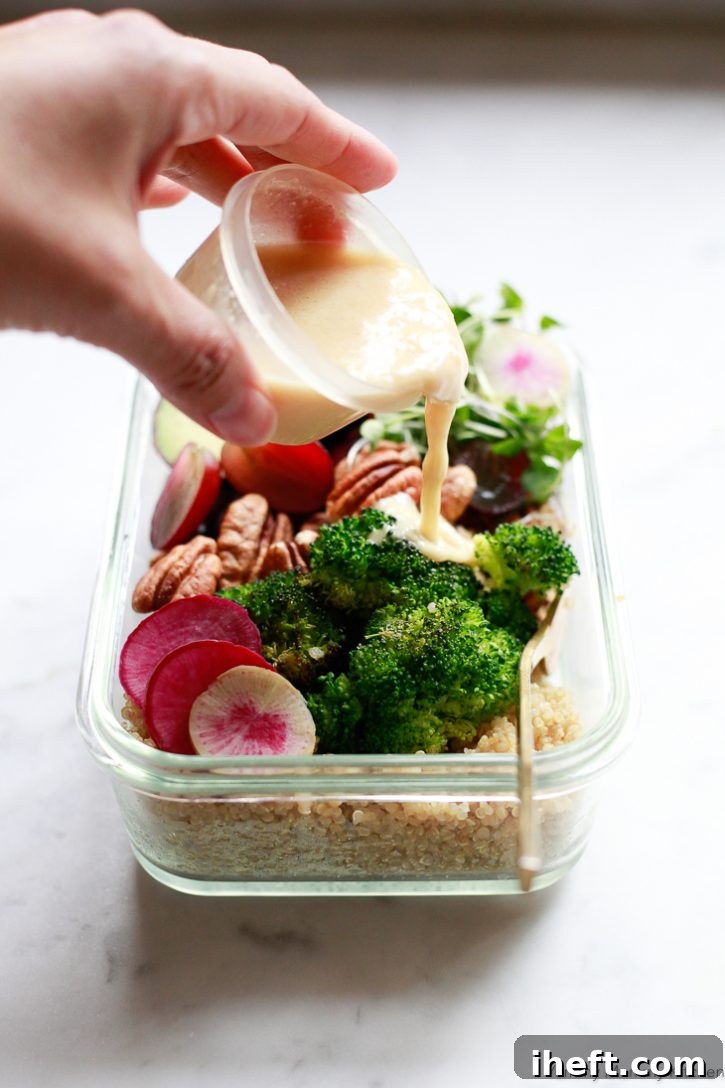 A hand gracefully drizzles a generous amount of homemade creamy lemon vinaigrette salad dressing over a vibrant vegan meal prep bowl, revealing the quinoa, roasted broccoli, and other fresh ingredients beneath.