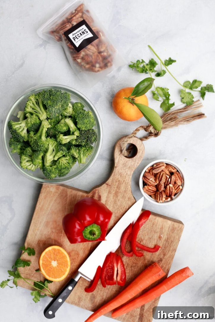 The ingredients for a vegetable stir fry sit on a white marble countertop and include broccoli, a red bell pepper, carrots, pecans, and an orange for the stir fry sauce.