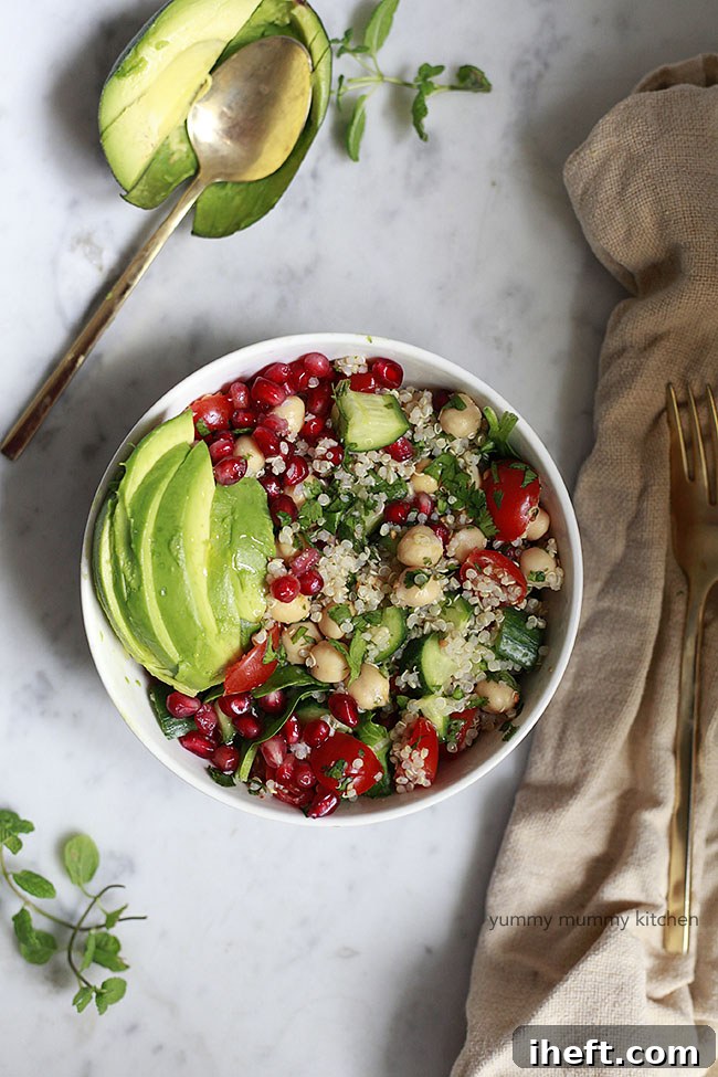 A white bowl filled with quinoa and chickpea tabbouleh, garnished with pomegranate arils and avocado slices, on a white marble countertop.