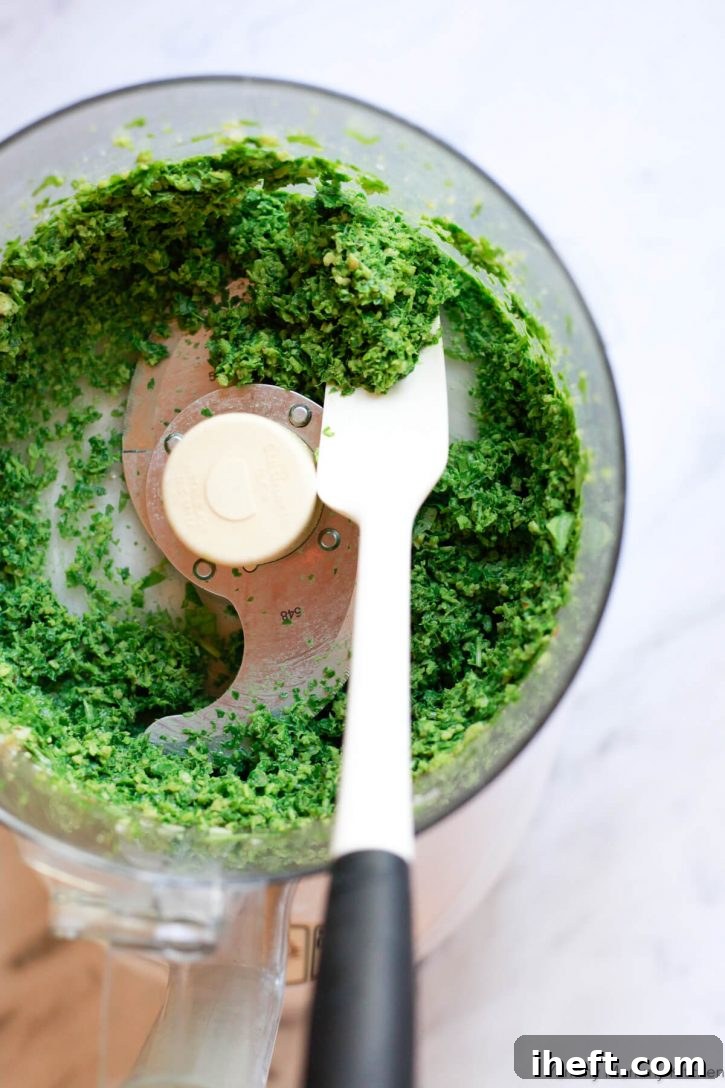 A white spatula scrapes down the sides of a food processor while making fresh basil pesto. 