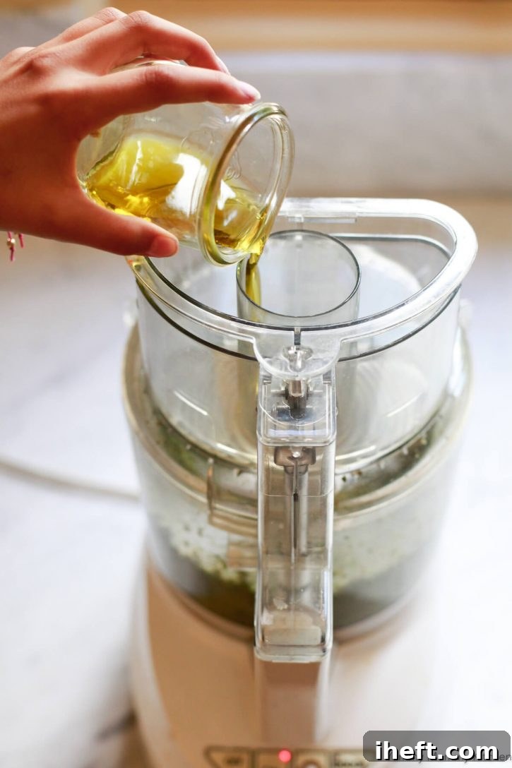 A hand pours a small jar of olive oil through the feed tube of a food processor while making vegan pesto. 