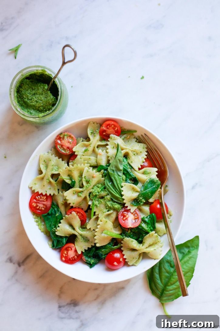 A bowl of vegan pesto pasta sits on a white marble countertop with a small jar of pesto sauce on the side. 
