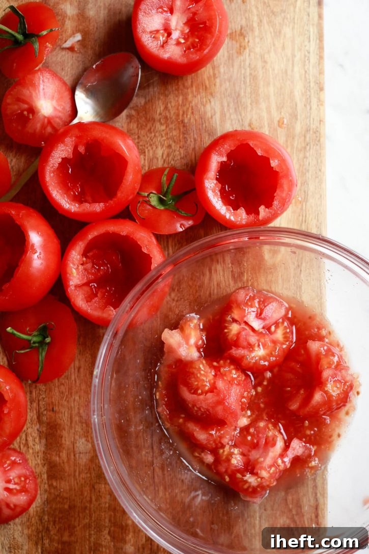 Tomatoes on tops cut off sit on a cutting board with pulp scooped out into a glass bowl. 