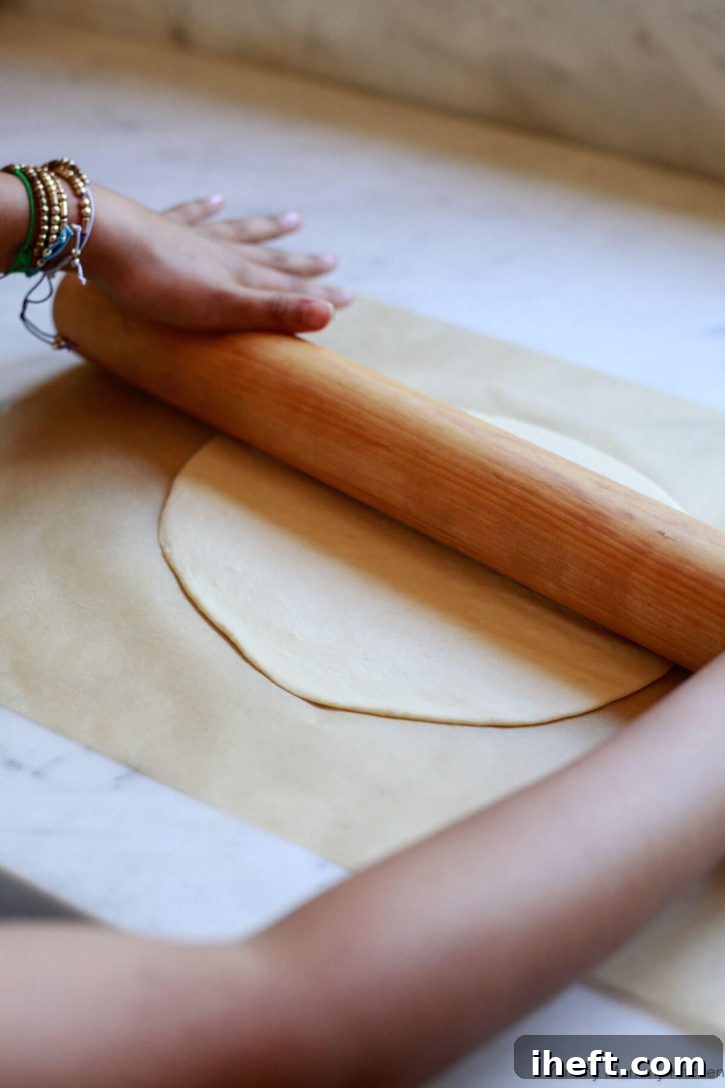 A child's hands roll a thin crust pizza dough with a wooden rolling pin. 