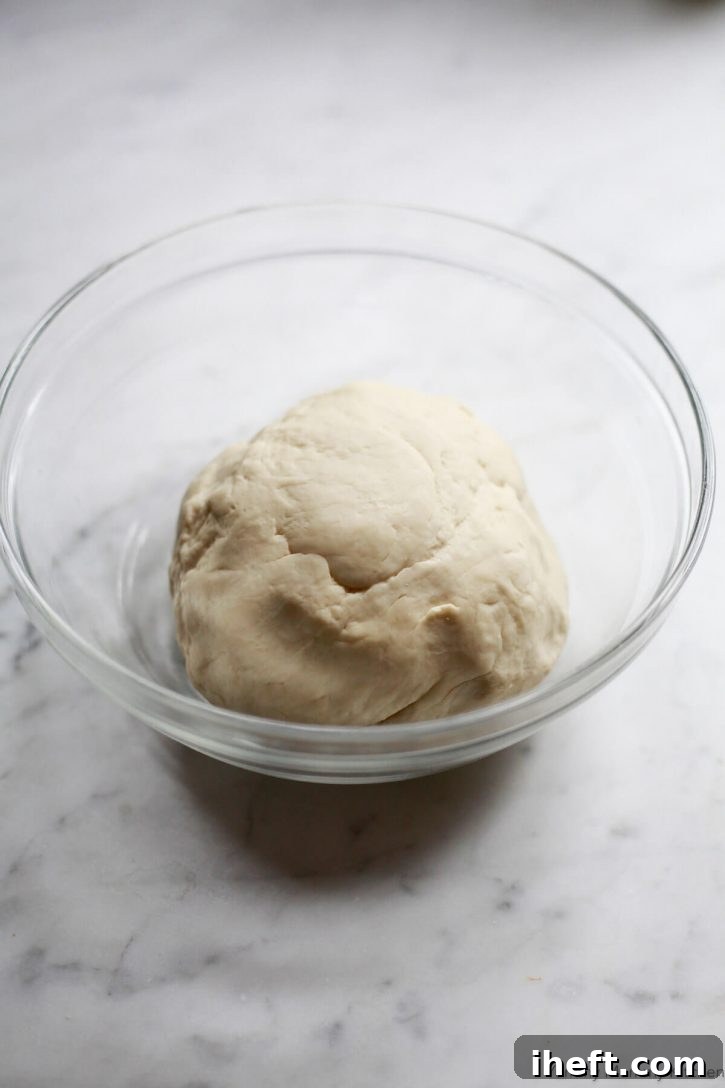 A ball of pizza dough sits in a glass bowl on a marble countertop to begin rising. 