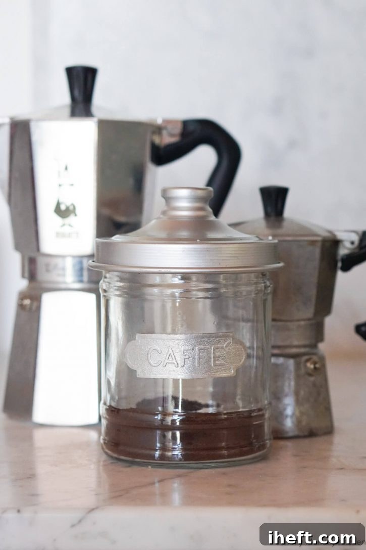 A glass canister of finely ground espresso coffee stands prominently in the foreground, with two traditional Bialetti Moka espresso makers subtly blurred in the background, illustrating the tools needed for authentic espresso.