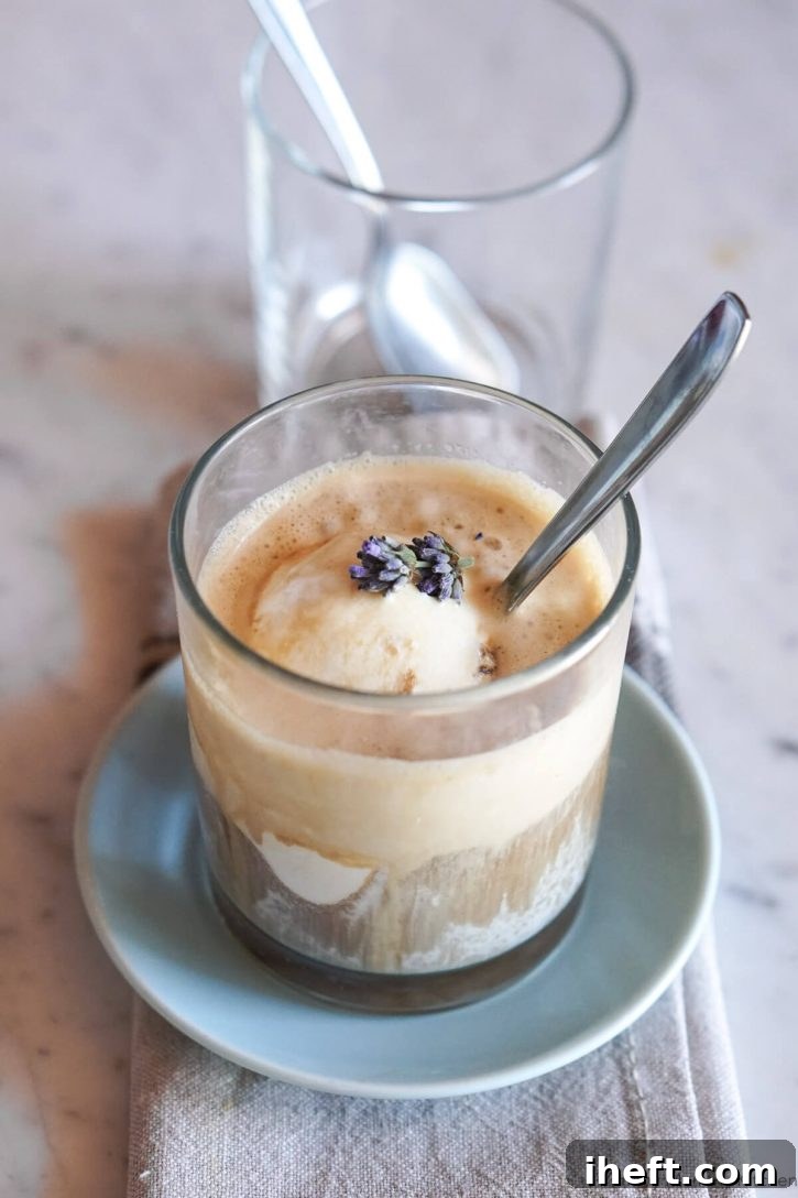 A perfectly prepared affogato, consisting of melted vanilla ice cream swirled with rich espresso, sits in an elegant glass with a slender metal spoon resting inside, all placed on a pristine marble countertop, highlighting this simple yet luxurious Italian coffee dessert.