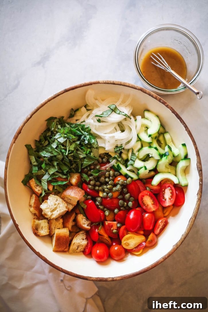 The ingredients for panzanella salad recipe are shown arranged in a white salad bowl. Tomatoes, cucumbers, onions, toasted bread, basil, and capers are pictured in this Italian tomato and bread salad with dressing in a jar on the side. This image shows how to make panzanella salad. 