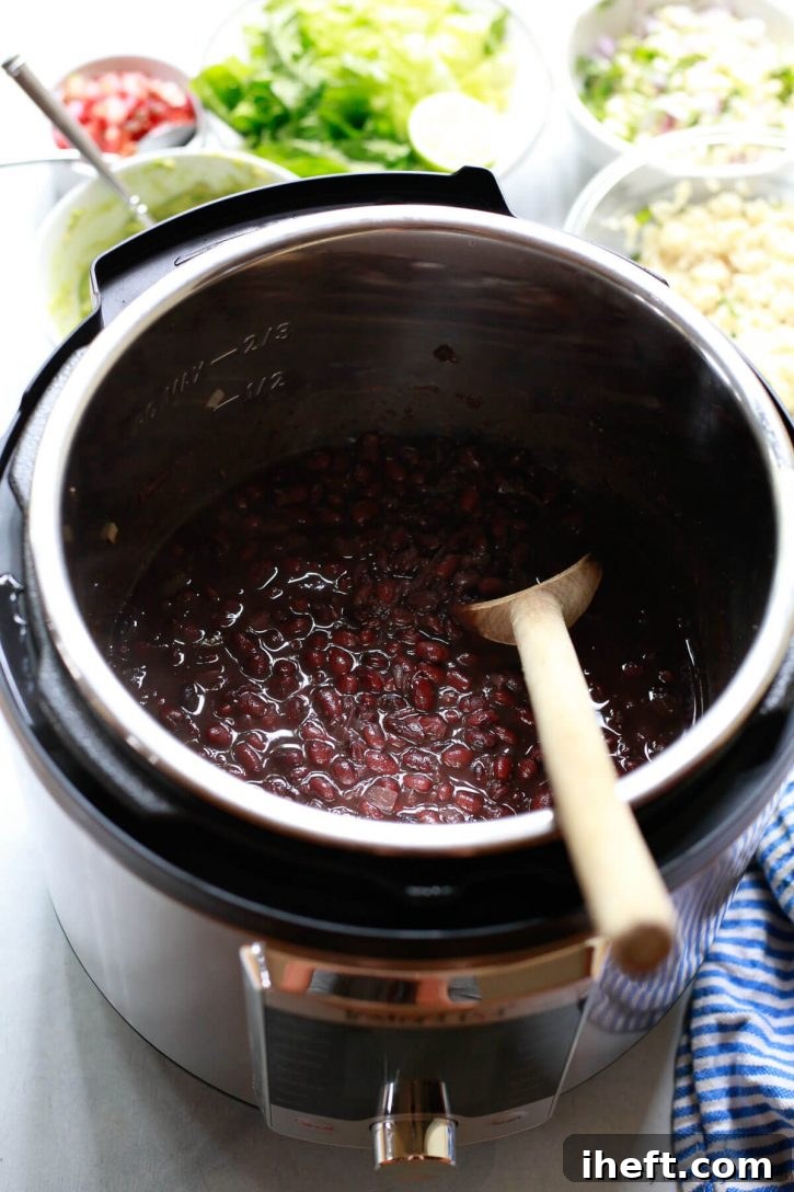 An Instant Pot pressure cooker filled with cooked black beans with burrito bowl toppings in the background. 