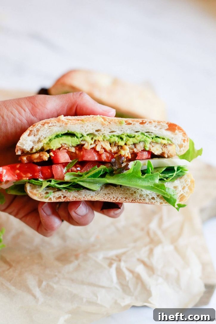 A hand holds a tempeh sandwich with tomato, lettuce, onion, and avocado. A great tempeh recipe!
