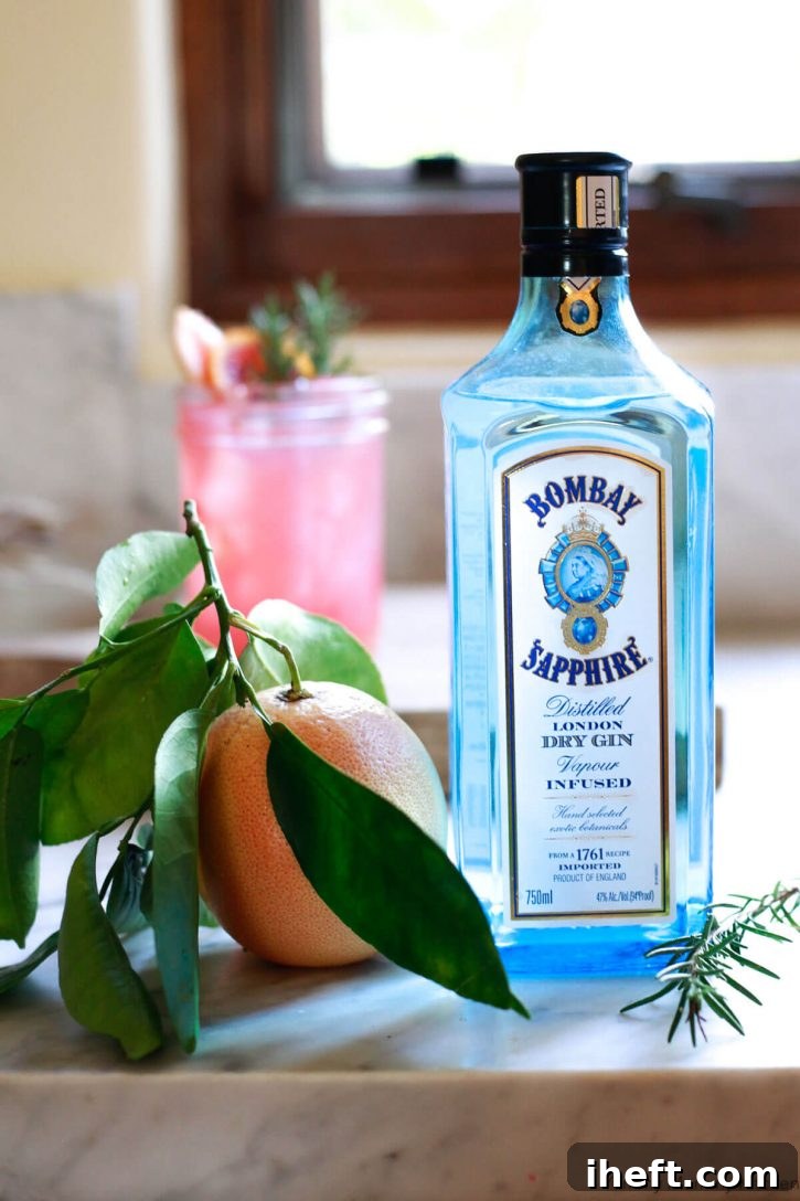 A bottle of Bombay Sapphire gin sits on a white countertop next to a fresh grapefruit with a greyhound drink in the background. 