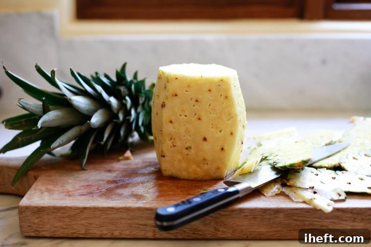 A whole fresh pineapple is peeled with a knife on a wooden cutting board. The initial steps of preparing a pineapple for slicing.