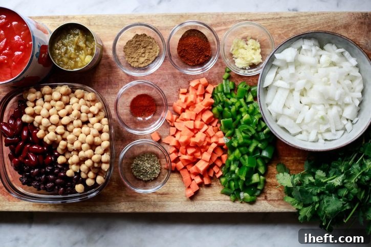 The ingredients for an easy vegan chili are set on a counter and include chickpeas, kidney beans, black beans, carrot, bell pepper, white onion, spices, green chilies, and diced tomatoes. 