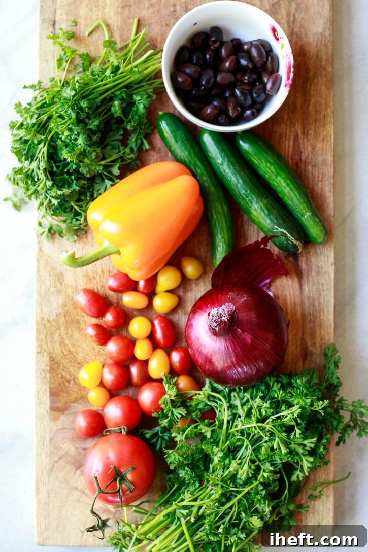 Fresh, vibrant Greek pasta salad ingredients including Kalamata olives, red onion, cucumber, cherry tomatoes, bell pepper, and fresh herbs arranged beautifully on a cutting board, ready for preparation.