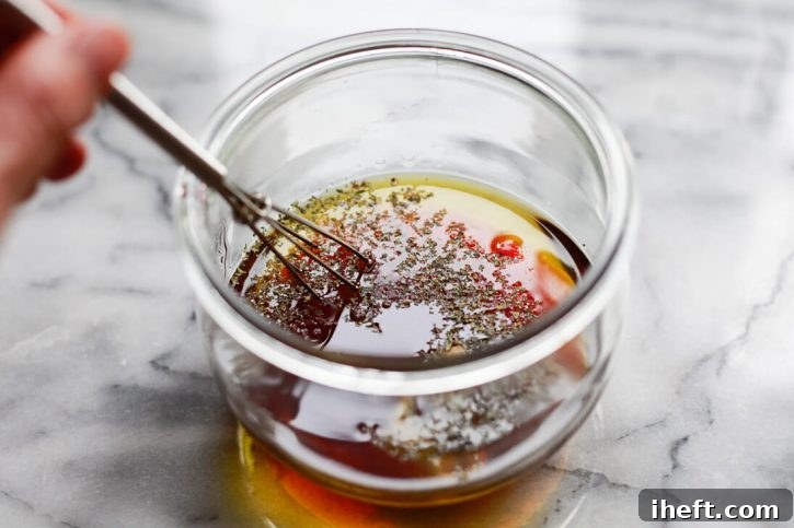 Olive oil, red wine vinegar, and dried oregano are being whisked together in a glass jar, creating a vibrant Greek-inspired pasta salad dressing.