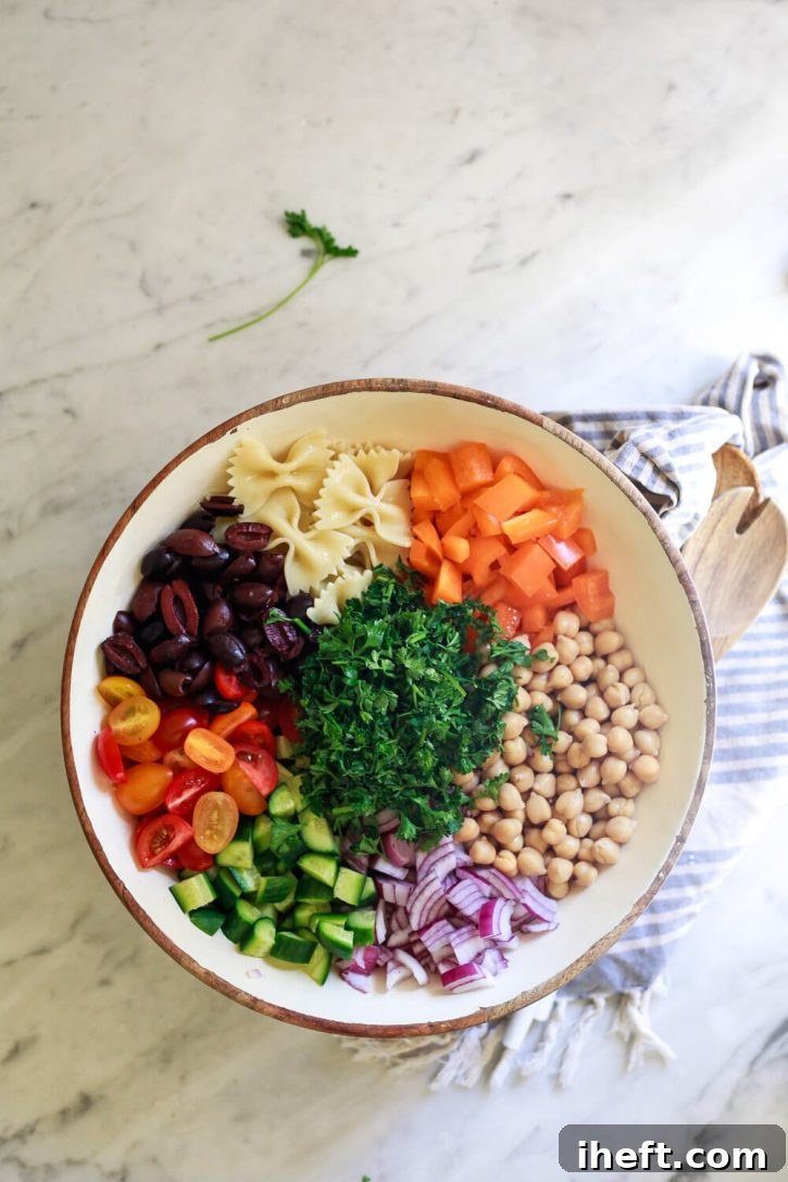A beautiful Greek pasta salad with bow tie pasta, orange bell pepper, chickpeas, red onion, cucumber, parsley, olives, and tomatoes, arranged in colorful sections in a large salad bowl on a marble countertop.
