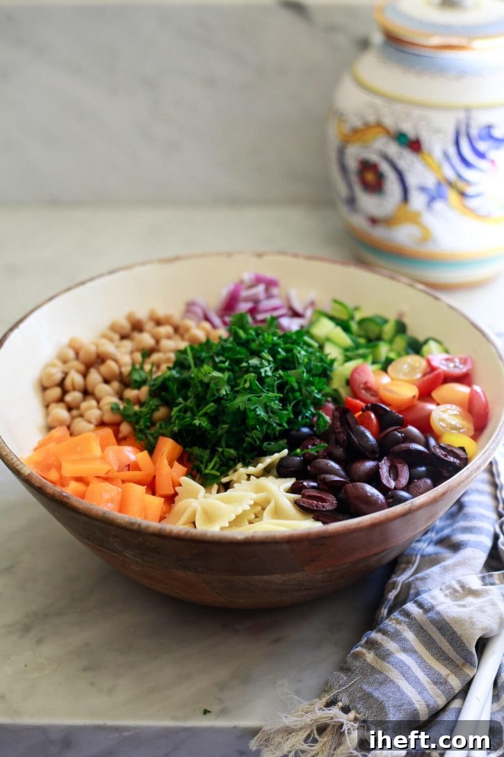 A wooden salad bowl filled to the brim with colorful ingredients for a Greek vegan pasta salad, showcasing the vibrant array of vegetables, pasta, and chickpeas.