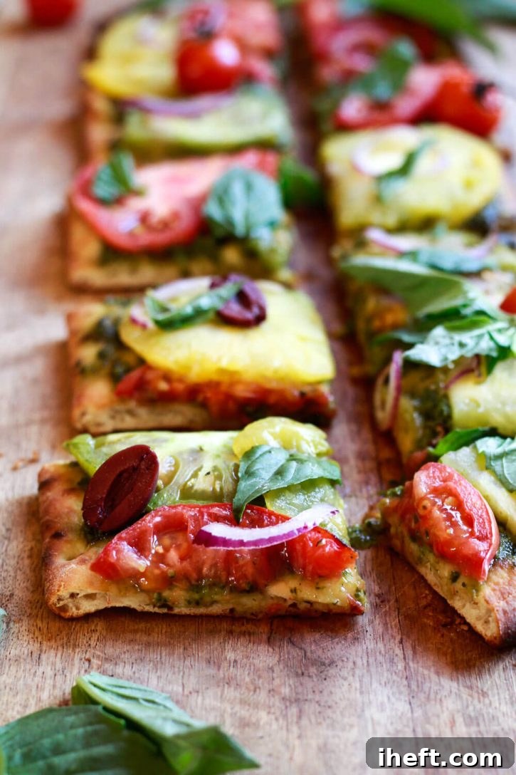 Slices of a delicious vegetarian and vegan pesto pizza generously topped with colorful heirloom tomato slices, fresh red onion, fragrant basil, and savory Kalamata olives, ready to be enjoyed.