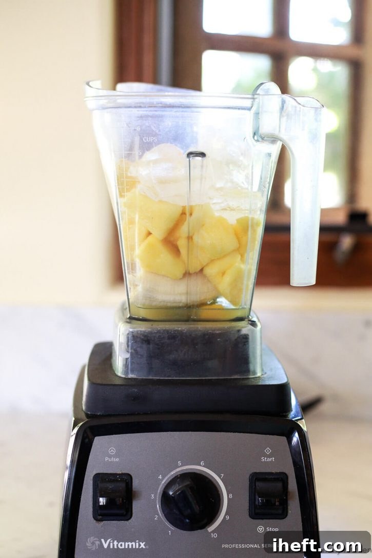 All the vibrant ingredients for a healthy vegan pineapple smoothie, including pineapple juice, peeled orange, banana, frozen pineapple, frozen mango, and ice, are perfectly layered in a Vitamix blender, ready for blending.