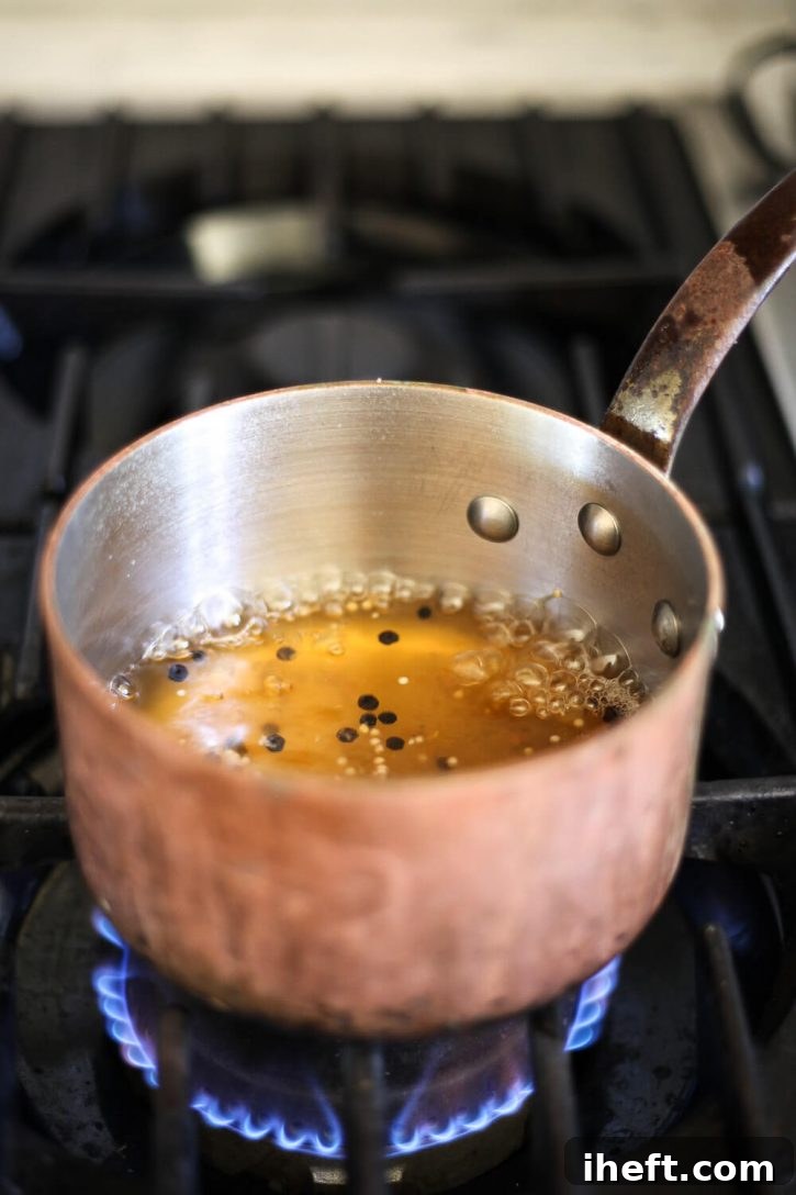 A copper saucepan on the stove containing apple cider vinegar, peppercorns, mustard seed, and brown sugar simmering to create the aromatic pickling liquid for beets.