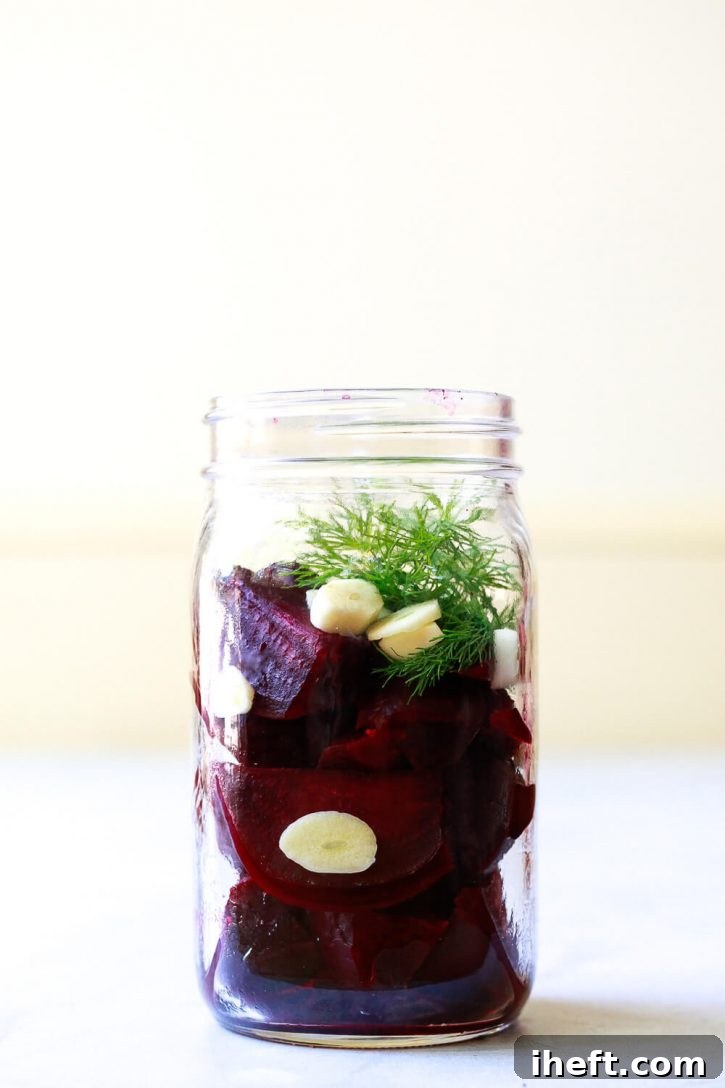 Clear pickling liquid being poured over prepared beet wedges, garlic slices, and fresh dill in a glass canning jar.