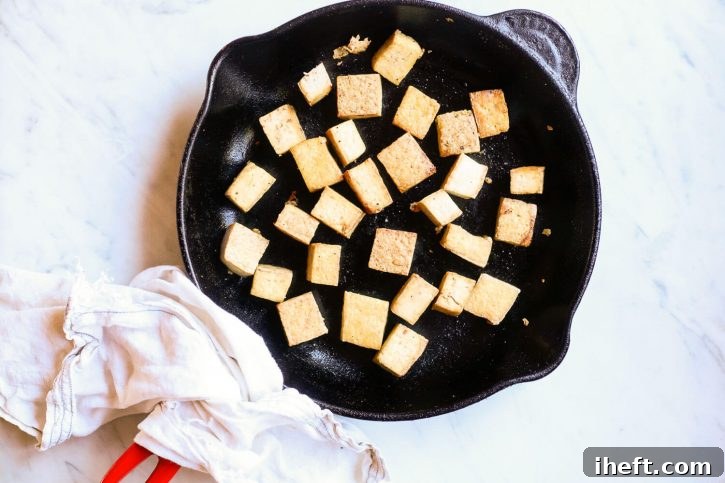Golden-brown, crispy pan-fried tofu cubes sizzling in a cast-iron Le Creuset skillet. A close-up shot showing the perfect texture achieved through pan-frying.