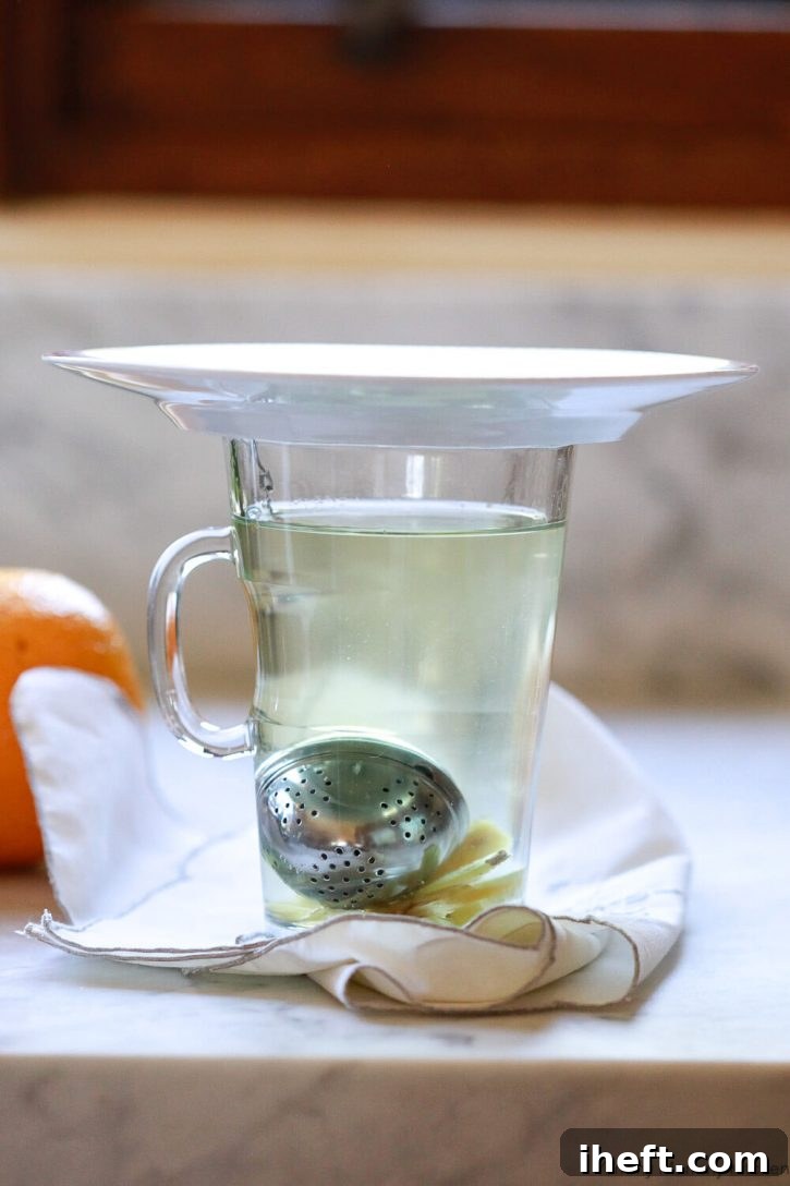 Homemade detox tea steeps in a glass tea cup with ginger at the bottom. The glass is covered with a small saucer to retain heat. 