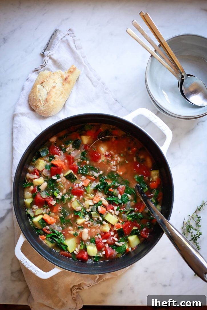 A white Staub cocotte pot filled with colorful, steaming Italian vegetable soup featuring white beans, kale, and farro, ready to serve.