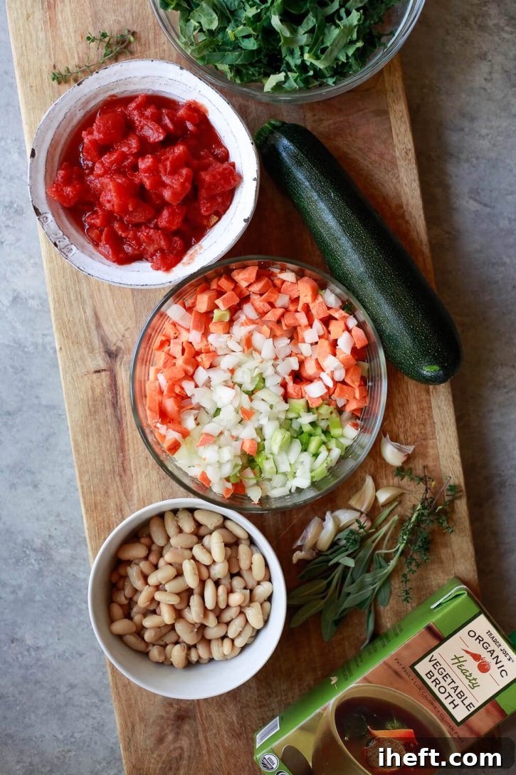 The vibrant ingredients for Italian vegetable soup laid out on a cutting board: neatly diced carrots, onion, and celery, fresh diced tomatoes, zucchini, creamy white beans, crisp kale, vegetable broth, and aromatic herbs.