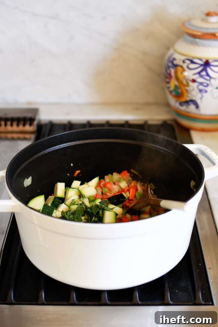 Fresh zucchini and fragrant herbs are carefully added to a simmering white Staub pot on the stove, enhancing a healthy vegetable soup recipe with vibrant color and flavor.