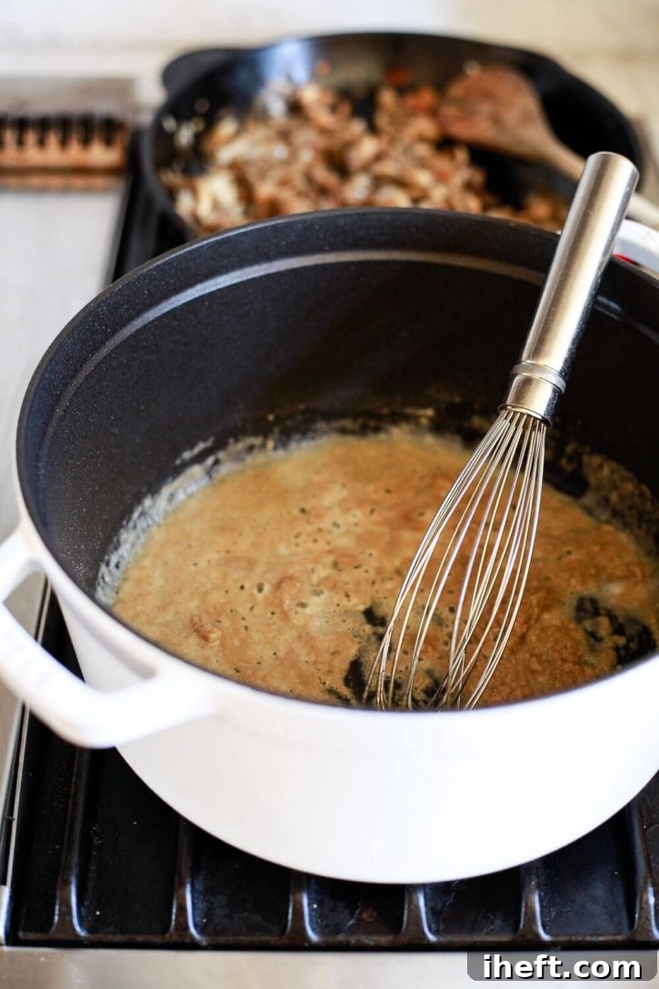 A whisk vigorously mixing flour into melted vegan butter and olive oil in a Staub pot, creating a light brown roux for the gravy.