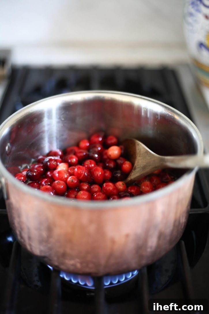 A close-up view of fresh cranberries simmering in a saucepan with orange juice and sweetener, some berries already starting to burst open. This indicates the sauce is perfectly cooking down to the desired consistency for homemade cranberry orange sauce.