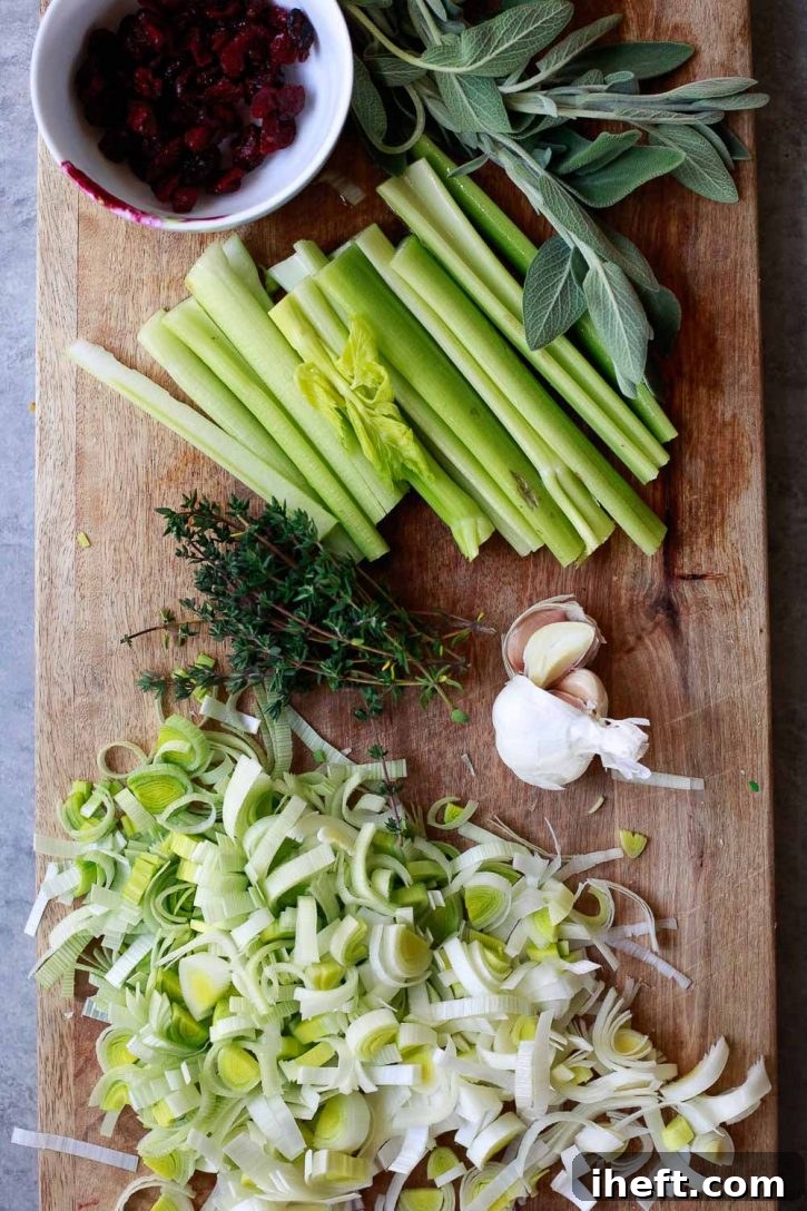 An inviting overhead view of classic stuffing ingredients neatly arranged on a rustic cutting board: vibrant green celery, fragrant leeks, fresh sprigs of thyme and sage, bright cloves of garlic, and ruby-red dried cranberries, all meticulously prepped for a wholesome vegan and vegetarian stuffing recipe.