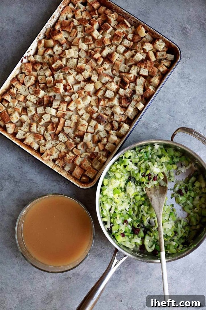 A beautifully composed flat lay showing the essential components for assembling vegan stuffing: a sheet pan filled with perfectly toasted golden-brown bread cubes ready for mixing, positioned next to a bowl of rich vegetable stock, and a skillet brimming with aromatic sautéed vegetables and herbs.