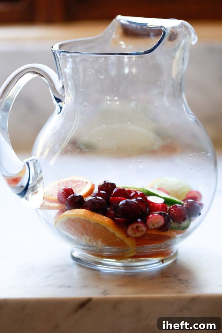 A large, clear glass pitcher showcasing a colorful medley of fresh fruit, including citrus slices and cranberries, settled at the bottom, ready to be infused for a delicious white sangria.
