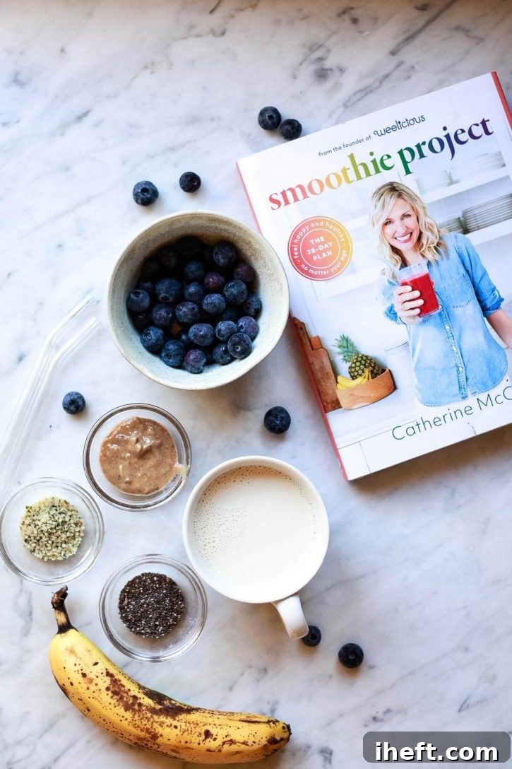 Fresh ingredients laid out on a marble countertop for a blueberry banana chia smoothie, alongside the 'Smoothie Project' cookbook. Ingredients include frozen blueberries, bananas, chia seeds, hemp seeds, almond butter, and a carton of almond milk.