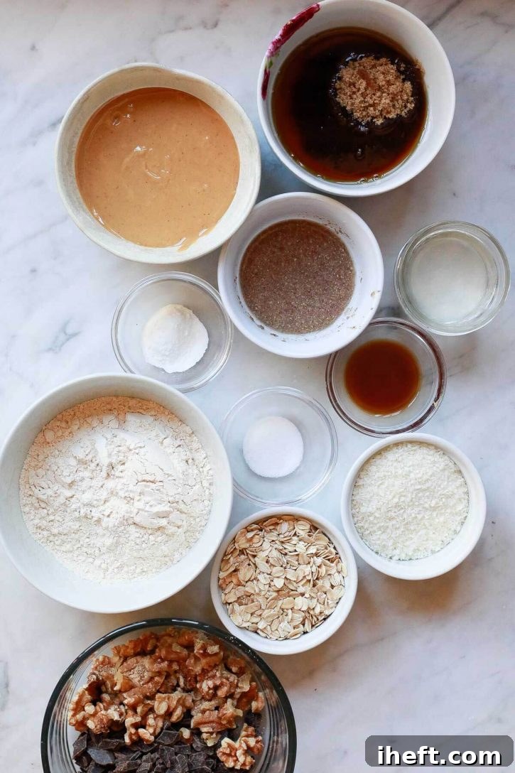 Bowls of essential cookie ingredients, including whole wheat pastry flour, rolled oats, creamy peanut butter, shredded coconut, and dark chocolate chunks, neatly arranged on a pristine marble countertop, ready for baking.