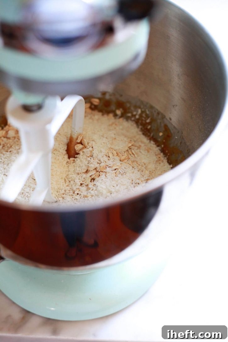 Dry cookie ingredients, including flour, oats, and coconut, being carefully added to the wet mixture in a KitchenAid mixer bowl, illustrating the next step in making vegan cookies.