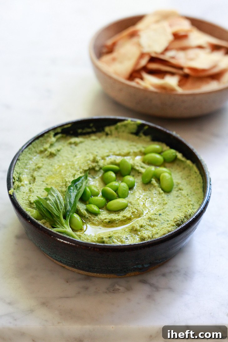 Bright green edamame hummus with fresh basil, served in a bowl with crunchy cucumber slices and pita bread.
