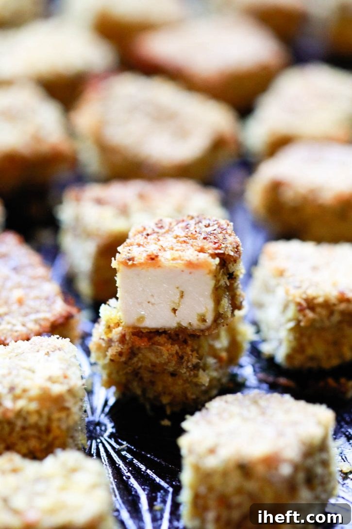 A close-up photo of crispy baked tofu nuggets on a cookie sheet. One tofu nugget is cut in half to reveal a juicy interior.