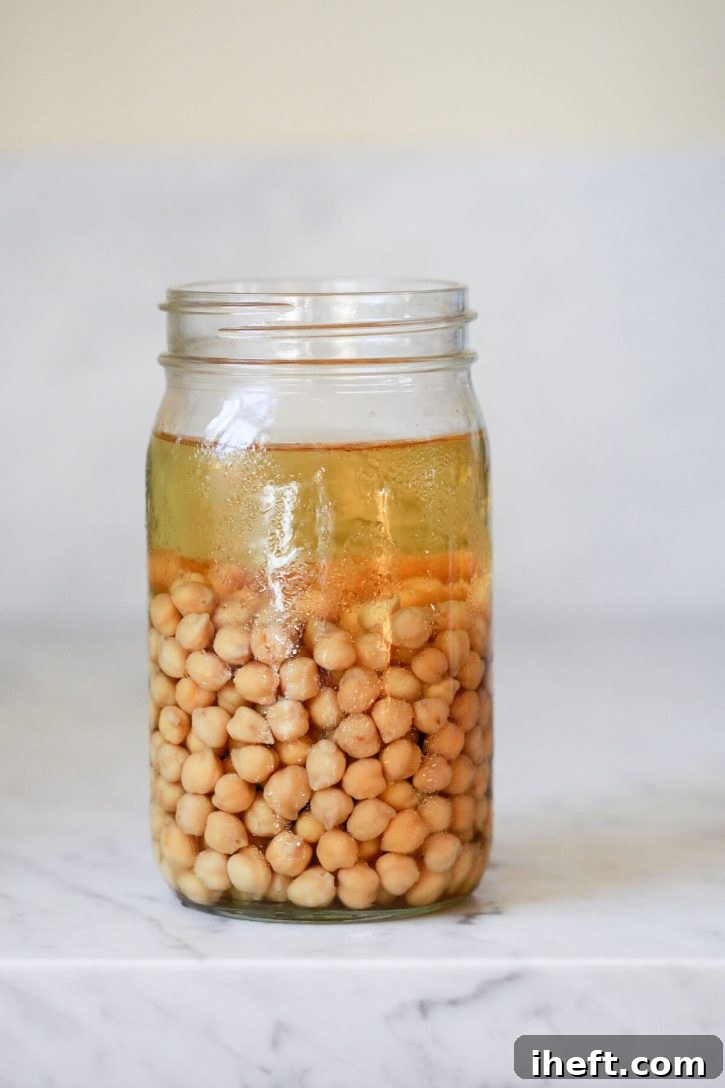 Dried chickpeas (garbanzo beans) soak in a glass jar on a white countertop, preparing for cooking.