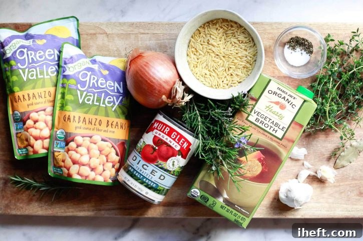 The ingredients for chickpea soup on a cutting board, including garbanzo beans, diced tomatoes, onion, broth, garlic, herbs, and orzo pasta. 