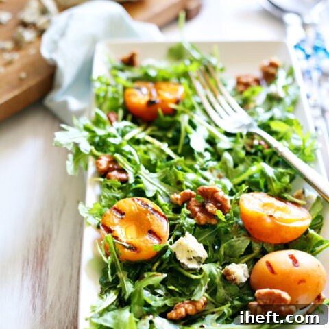 A summer salad of arugula, grilled apricots, gorgonzola, and nuts on a white plate.