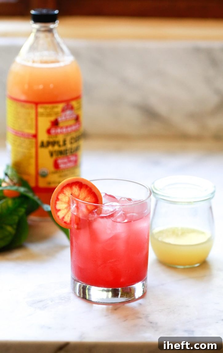A tall glass filled with water, beautifully mixed with a blood orange apple cider vinegar shot, creating a vibrant, healthful beverage. A classic bottle of Bragg's organic ACV with the mother stands clearly visible in the background, signifying the quality ingredient used.