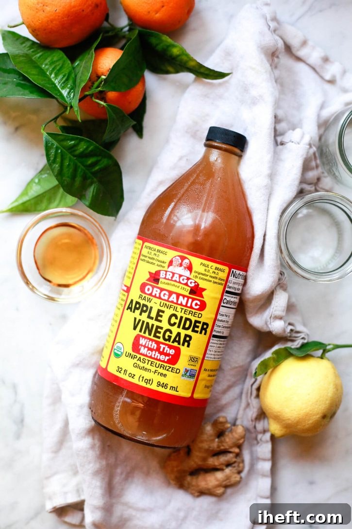 A clean, minimalist shot of the key ingredients for an invigorating apple cider vinegar shot recipe laid out on a shimmering marble counter. Featured prominently are a bottle of Bragg's apple cider vinegar with the mother, a knob of fresh ginger, and bright yellow lemons, signaling purity and natural efficacy.