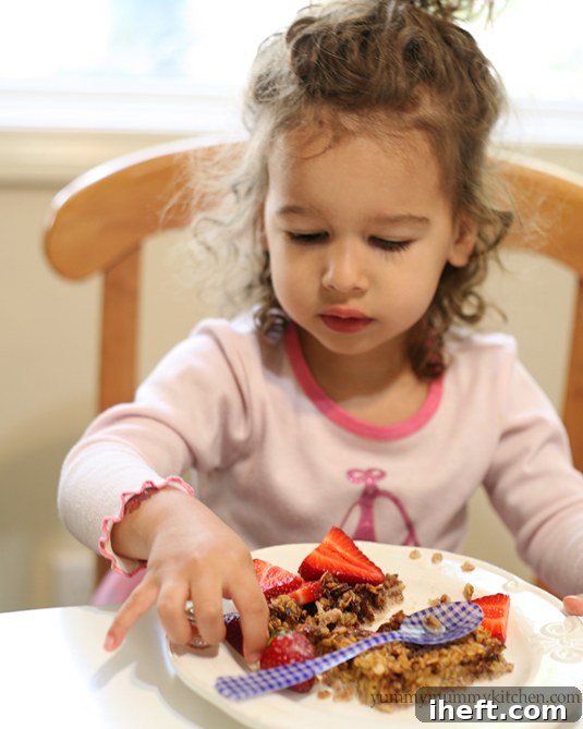 A toddler girl in a pink shirt eats a piece of baked oatmeal with berries.