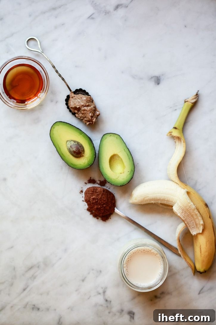 The ingredients for a chocolate banana avocado smoothie on a marble kitchen counter: banana, avocado, cacao powder, almond milk, almond butter, and maple syrup. 