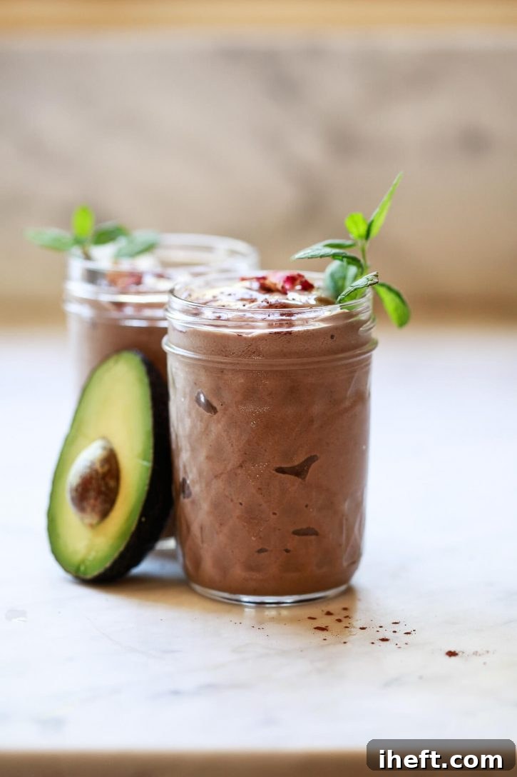 A chocolate banana avocado smoothie on a marble counter sitting next to half of an avocado. A healthy vegan chocolate avocado smoothie recipe. 