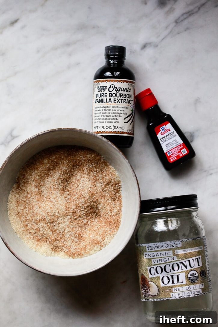 A bowl of sugar, jar of coconut oil, and bottles of vanilla and coconut extract neatly arranged on a marble counter, showcasing the simple ingredients needed for a DIY sugar scrub recipe.