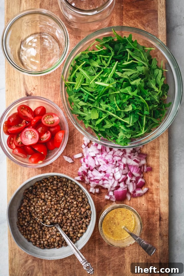 Cold French lentil salad ingredients sit on a cutting board to assemble into mason jars. Ingredients shown include cooked French lentils, halved cherry tomatoes, arugula, diced red onion, and a prepared homemade mustard vinaigrette. 